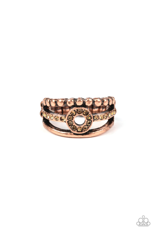 A smooth and dotted copper band flank a topaz rhinestone encrusted band, creating glittery layers across the finger. Encrusted in golden topaz rhinestones, a circular frame is pressed into the center of the bands for a refined finish. Features a dainty stretchy band for a flexible fit.