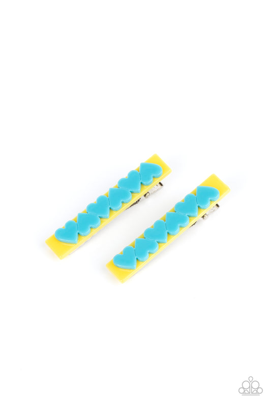 A row of blue hearts alternate across the front of a yellow rectangular frame, creating a charming pair of flirtatious hair clips. Features standard hair clips on the back.  Sold as one pair of hair clips.