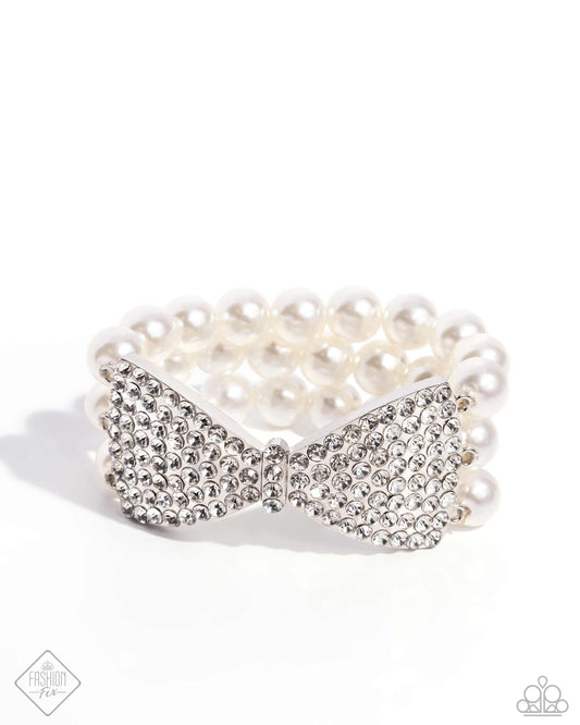 Strung along elastic stretchy bands, three bands of glossy white pearls wrap around the wrist. Featured in the center of the pearly collection, a white rhinestone-embellished silver bow flares out, creating a dapper stacked statement.  Sold as one individual bracelet.