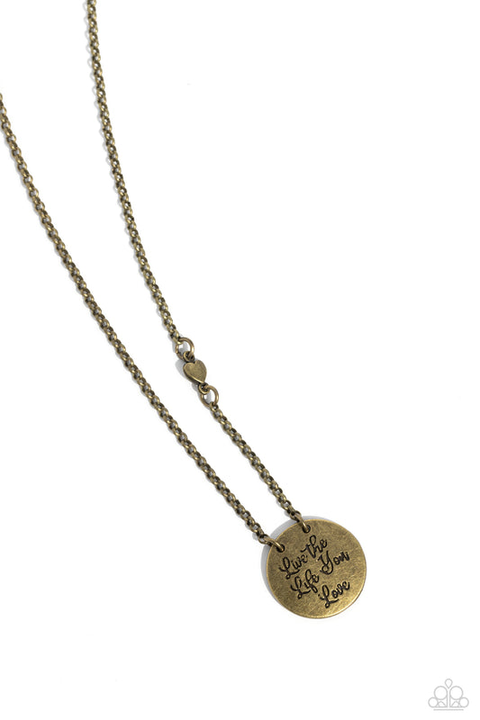A burnished brass disc is stamped in the inspiring phrase, "Live the Life You Love," at the bottom of a dainty brass chain, creating a motivational pendant. A dainty brass heart bead adorns one side of the chain for a light-hearted finish. Features an adjustable clasp closure.  Sold as one individual necklace. Includes one pair of matching earrings.